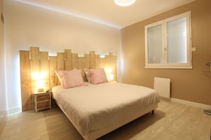 Un dormitorio con cabecera de madera, cama con ropa blanca, mesita de noche con lámpara y ventana con persianas.