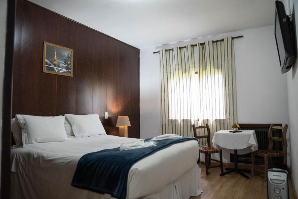 Quarto de hotel com cama, mesa, cadeiras, televisão e janela com cortinas.
