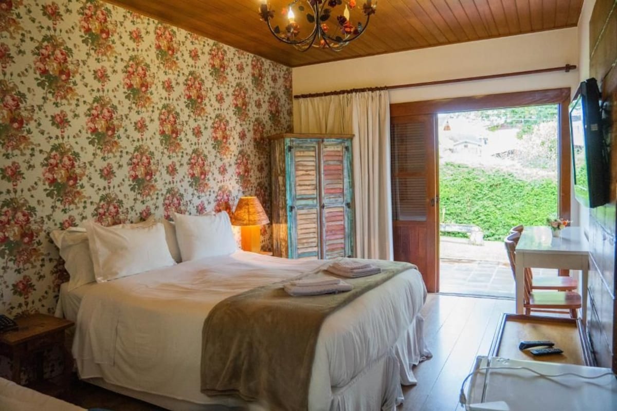 Quarto com cama, mesa de cabeceira, luminária, cadeira de madeira e vista para o exterior através de uma porta.