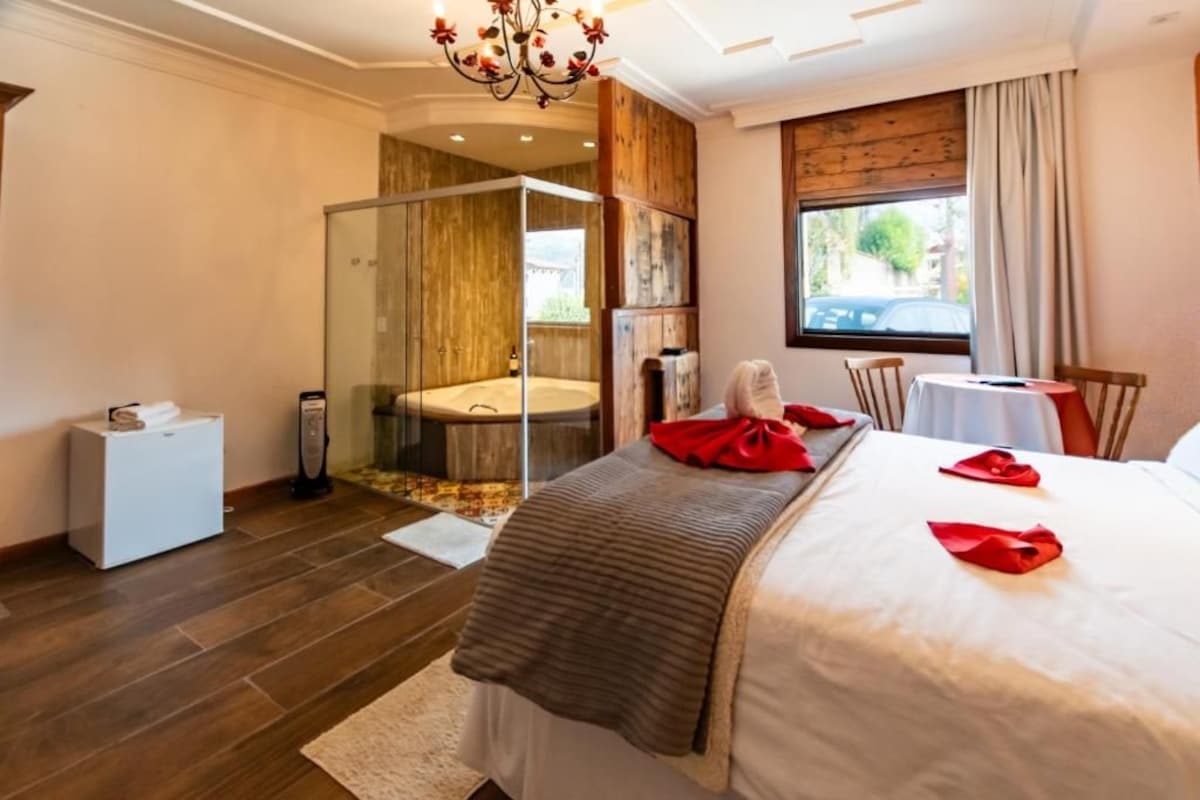 Quarto de hotel com cama, chuveiro em box, uma mesinha e uma janela.