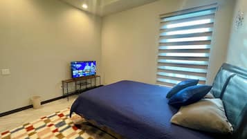 A bedroom with a bed, a television on a stand, a window with blinds, and a patterned rug.
