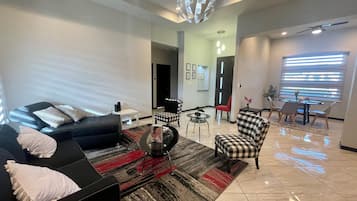 A modern living room with a black sofa, a checkered ottoman, a glass coffee table, and a dining area with a table and chairs.