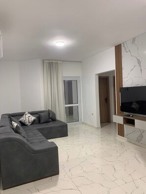 A modern living room with a sectional sofa, a flat-screen TV, and a sliding glass door leading to a balcony. - La Prestigia 2BR With a big Balcony (Hurghada)