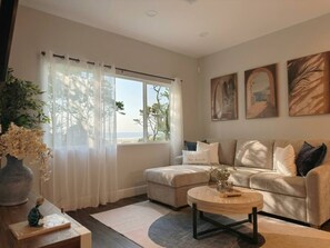 A living room with a sofa, ottoman, coffee table, and a view of the outdoors through a window with sheer curtains.