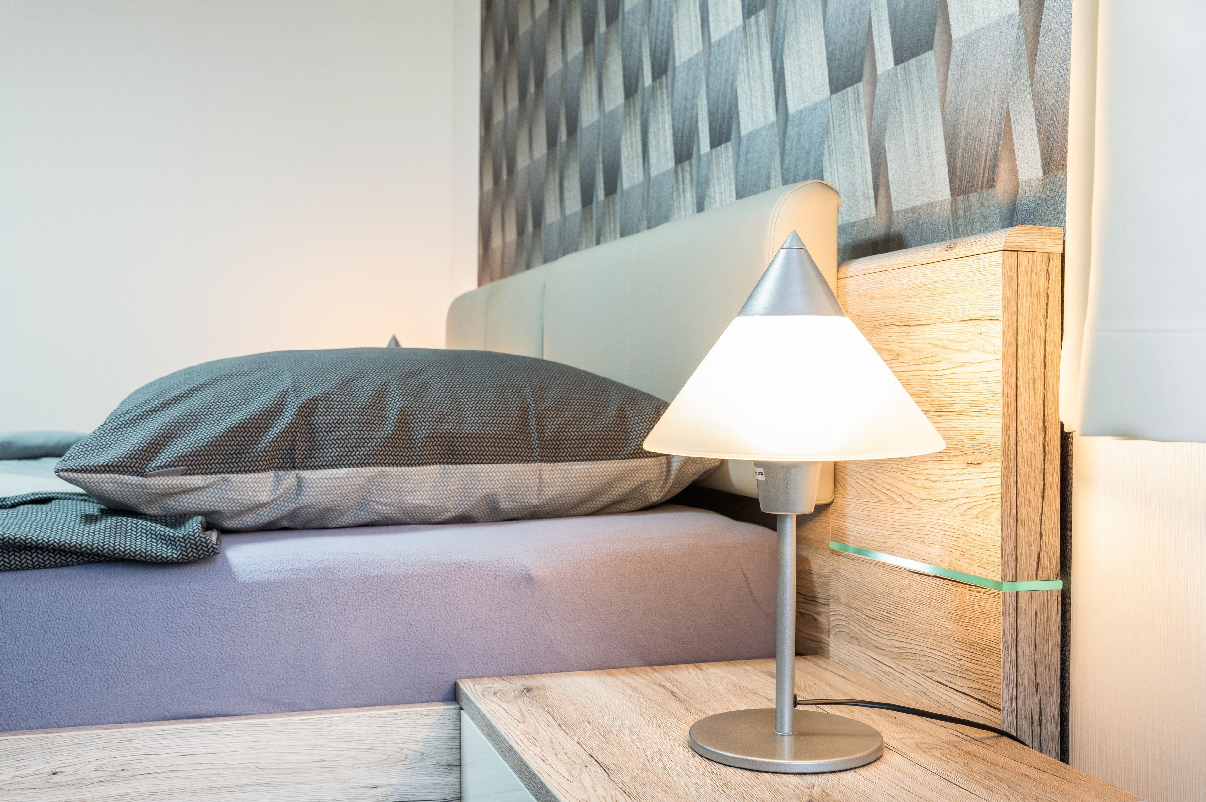 Et moderne soveværelse med seng, natbord, lampe og et træpanel bag hovedgæret.