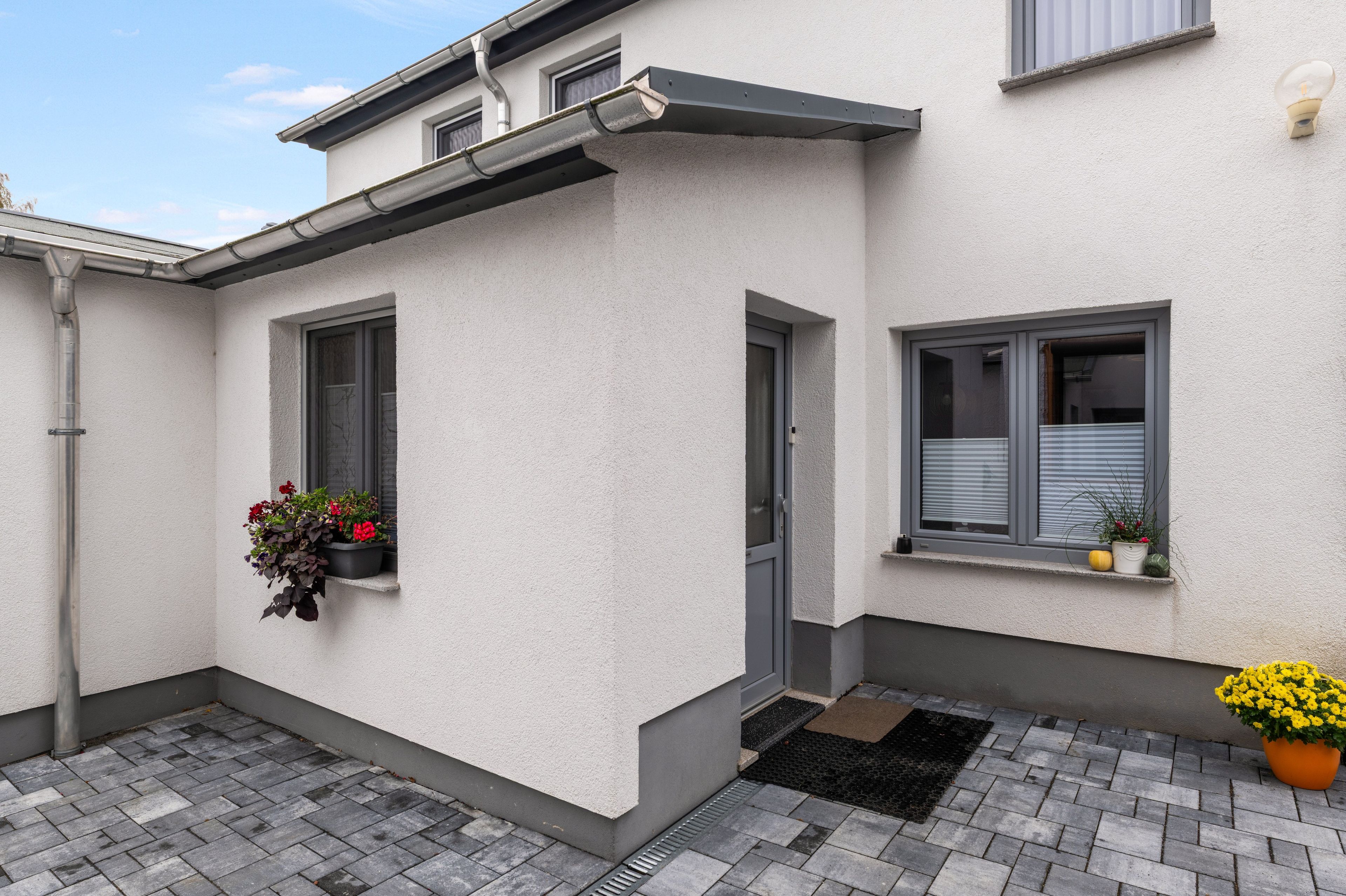 Ein Gebäude mit weißer Fassade, grauen Fensterrahmen, einer Tür und einem kleinen Fenster mit Pflanze.