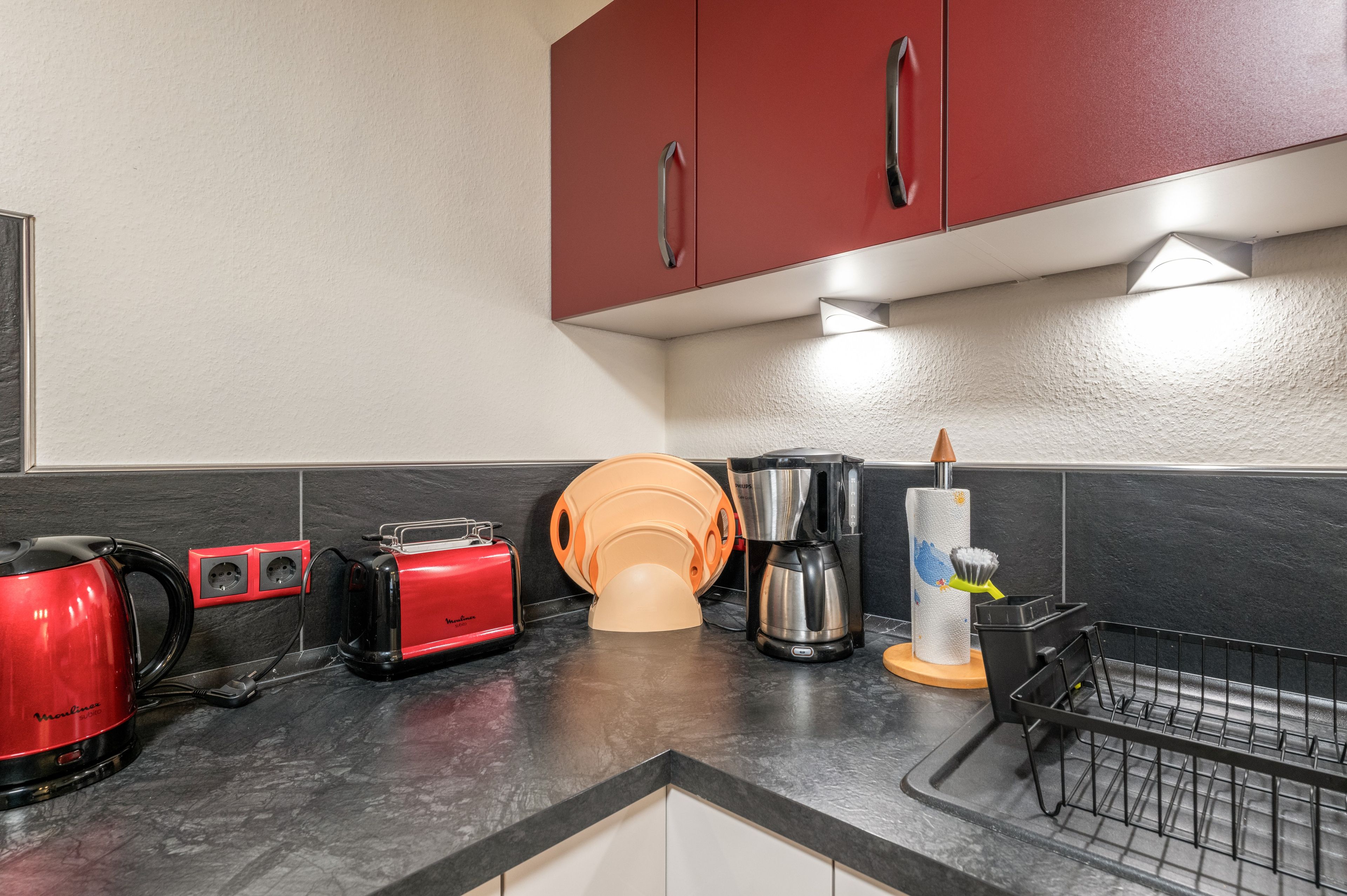 Eine Küche mit einem roten Toaster, einer Kaffeemaschine und einem Wasserkocher auf einer schwarzen Arbeitsfläche.