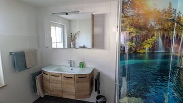 Ein modernes Badezimmer mit großem Spiegel, Holzwaschtisch und einer Dusche mit Glastrennwand und Blick auf einen Wasserfall.