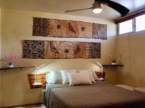 Quarto com cama, duas luminárias na parede, ventilador de teto e duas decorações penduradas.