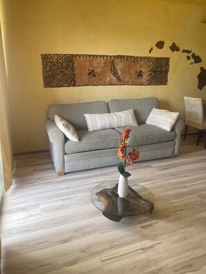 A living room with a grey sofa, a wooden coffee table with a vase of flowers, and a wall tapestry. - Cozy 1-bedroom condo in charming Paia with AC in the bedroom. (Paia)