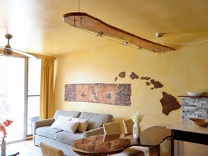 A modern living room with a sofa, a coffee table, a dining area with a bowl, and a ceiling fan. - Cozy 1-bedroom condo in charming Paia with AC in the bedroom. (Paia)