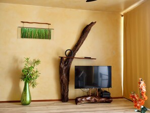 Quarto com televisão, escultura de madeira, planta verde em vaso e piso de madeira.