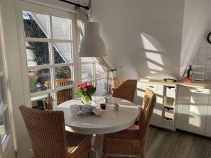 A kitchen with a round table, wicker chairs, a hanging light fixture, and a view through large windows. - Holiday Apartment in Nieblum With Balcony (Nieblum)