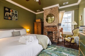 A hotel room with a large bed, a brick fireplace, a round mirror, a window with curtains, and a painting on the wall. - Gertrude: 1882 Romantic Victorian, 2 Kings, Private Hot Tub, near Forsyth Park (Savannah)
