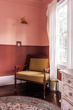 A room with a wooden chair, a small table, a decorative lamp, and a window with curtains. - Gertrude: 1882 Romantic Victorian, 2 Kings, Private Hot Tub, near Forsyth Park (Savannah)