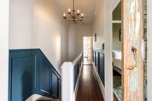 A hallway with a chandelier, a staircase with blue railings, and a bed in the adjacent room. - Gertrude: 1882 Romantic Victorian, 2 Kings, Private Hot Tub, near Forsyth Park (Savannah)