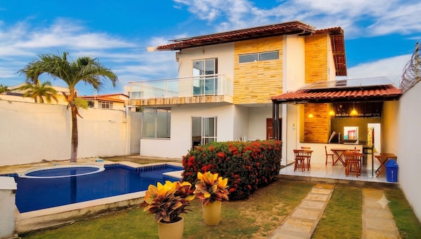 A two-story house with a swimming pool, a patio with outdoor furniture, and a small garden with colorful flowers.