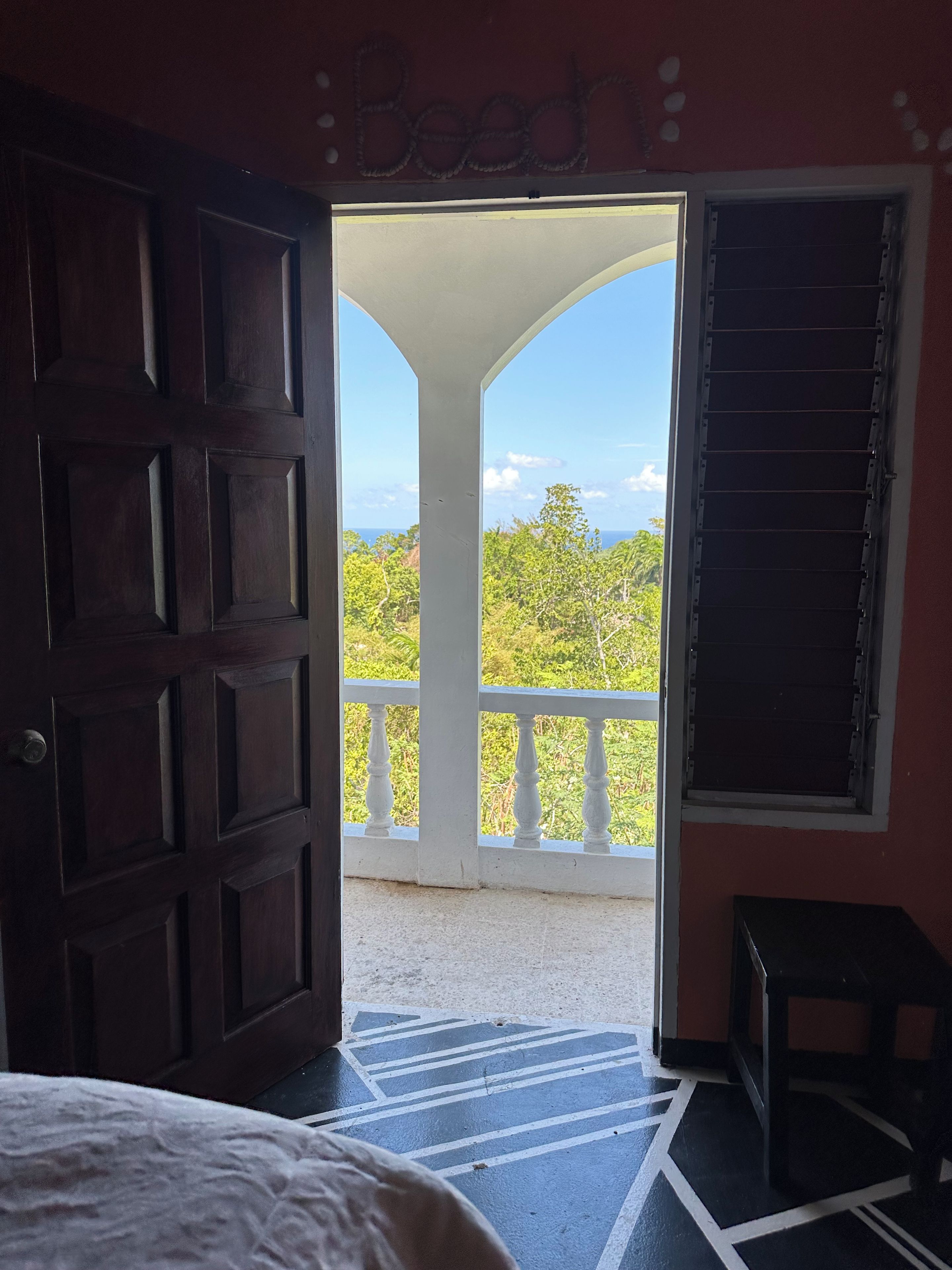Une pièce dotée d’une grande porte en bois, d’un balcon avec une balustrade blanche et offrant une vue sur la verdure et le ciel.