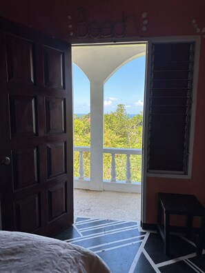 A room with a large wooden door, a balcony with a white railing, and a view of greenery and the sky.