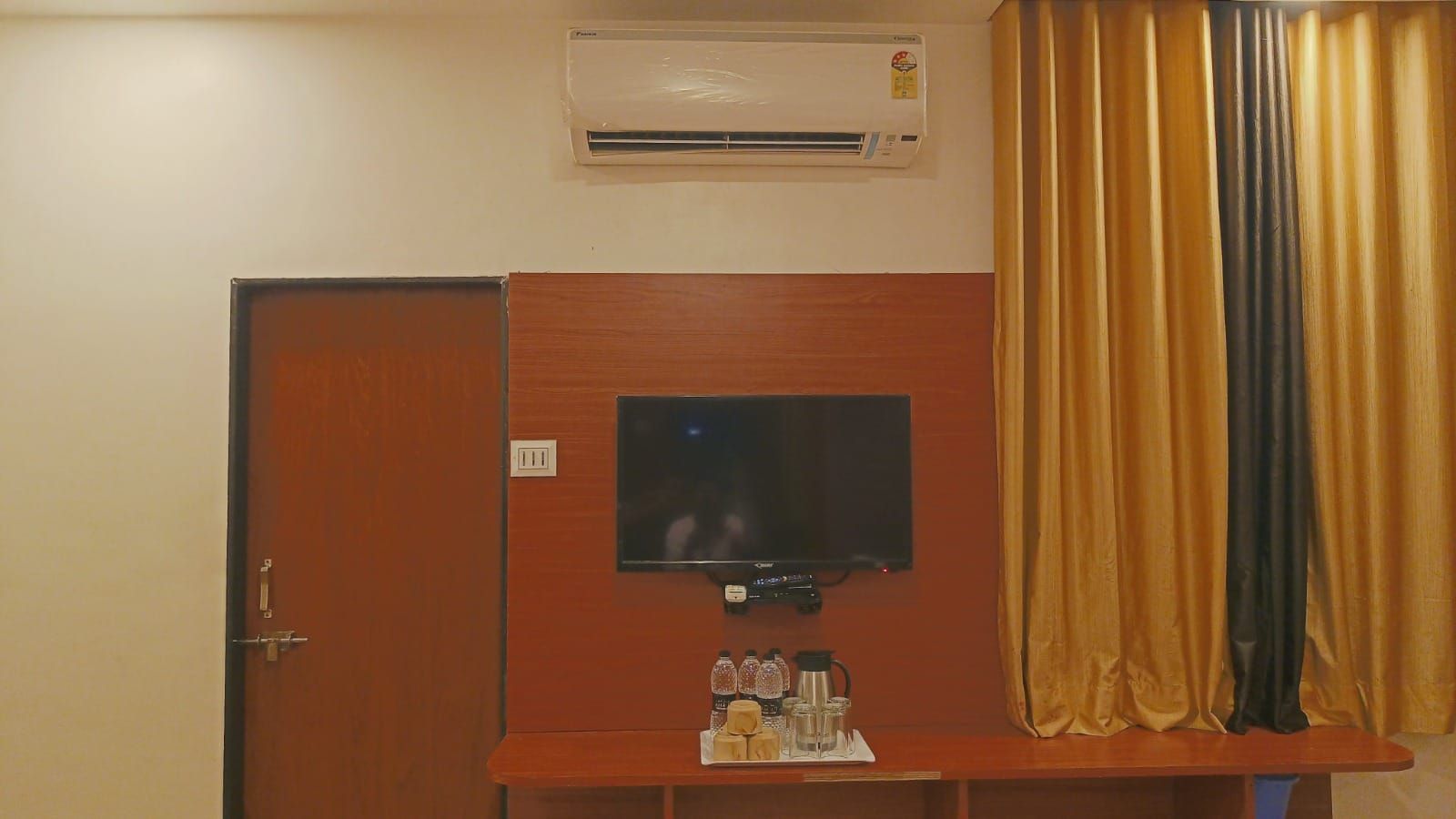 A room with a closed door, an air conditioning unit, a flat-screen TV mounted on a wooden panel, and a curtain.