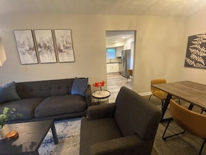 A living room with a sofa, a coffee table, and a dining table with chairs. There is a kitchen in the background. - Updated 3-bedrm house in adorable Stone Mtn area! Near to all Atlanta's venues! (Snellville)