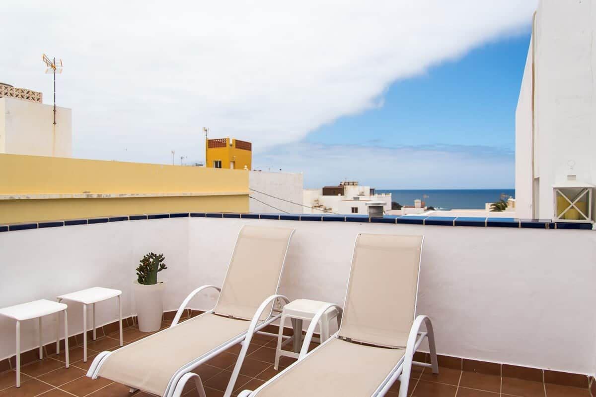 A rooftop terrace with lounge chairs and small tables, overlooking a coastal cityscape.