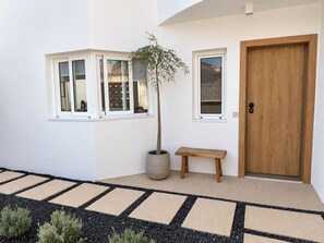 A building exterior with a wooden door, a bench, a potted plant, and a window with a view of a building. - Spherae Magma (Corralejo)