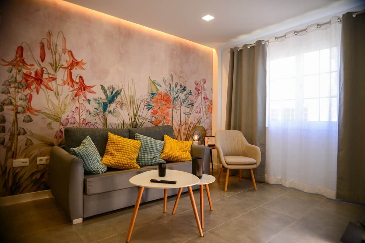 A living room with a sofa, a small round table, a chair, and a floral wall mural.