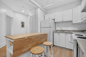 Una cuina moderna amb armaris blancs, una illa de fusta amb tamborets, fogó integrat i frigorífic.