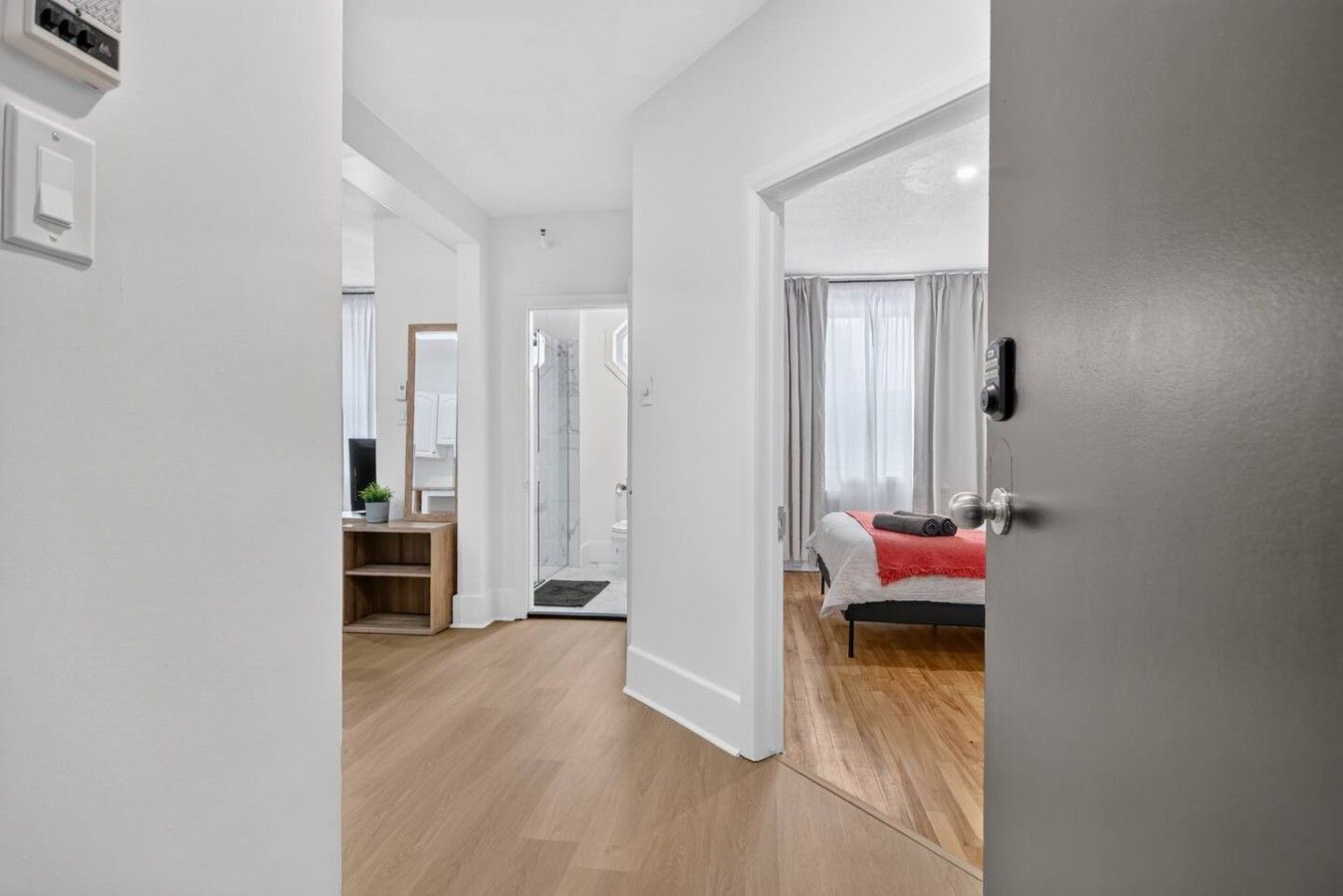 Une chambre d’hôtel moderne avec un plancher en bois, un lit recouvert d’une couverture rouge, une salle de bain avec douche et un bureau équipé d’un ordinateur.