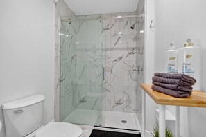 A bathroom with a glass shower enclosure, a toilet, and a shelf with folded towels and Dove skincare products. - The Modern Corner 1BR | Kitchen, A/C & Wi-Fi (Gatineau)