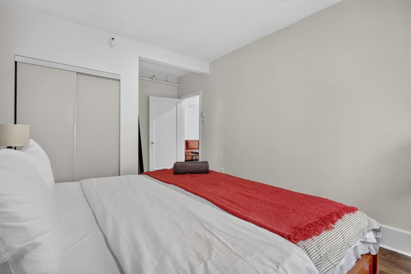 Een slaapkamer met een bed, een rode plaid, een stoel en een deur die naar een andere kamer leidt.