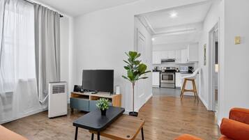 A modern living room with a flat-screen TV, a coffee table, and a sofa. A kitchen is visible in the background with white cabinets and a stove.