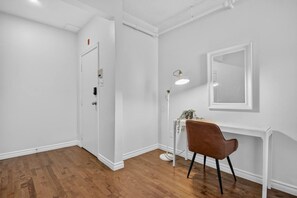 A room with a wooden floor, white walls, a door, a desk with a chair, and a floor lamp. - Capital Region 1BR | Minutes to Attractions (Gatineau)