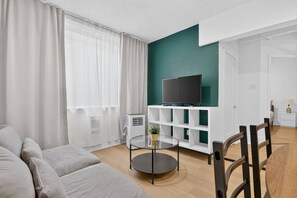 A modern living room with a sofa, a glass coffee table, a TV on a white shelf, and a small plant. - Modern 1BR Oasis | Steps to City Highlights (Gatineau)