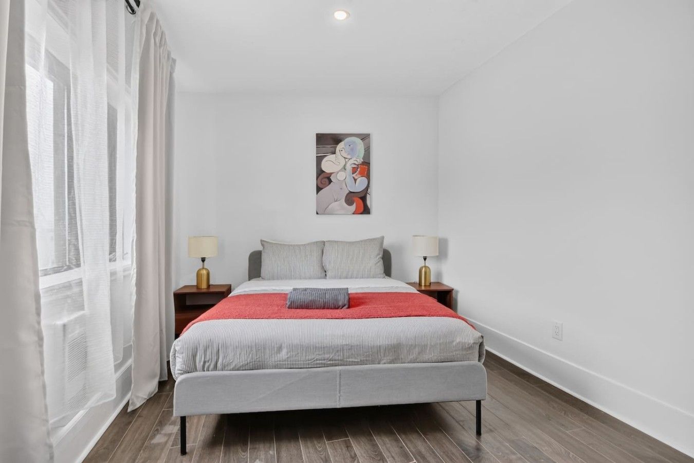 A bedroom with a bed, two bedside tables, a window with sheer curtains, and a wall-mounted artwork.