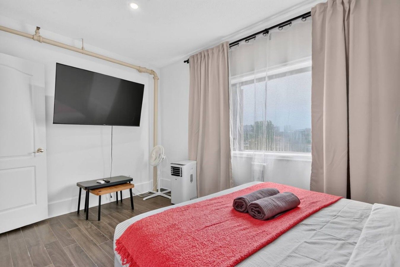 Une chambre d’hôtel comprenant un lit, un téléviseur fixé au mur, un bureau, une chaise, une fenêtre avec des rideaux et un appareil de climatisation.