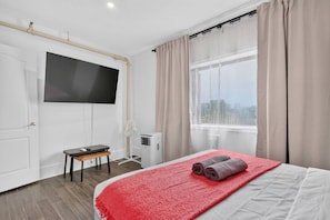 Une chambre d’hôtel comprenant un lit, un téléviseur fixé au mur, un bureau, une chaise, une fenêtre avec des rideaux et un appareil de climatisation.