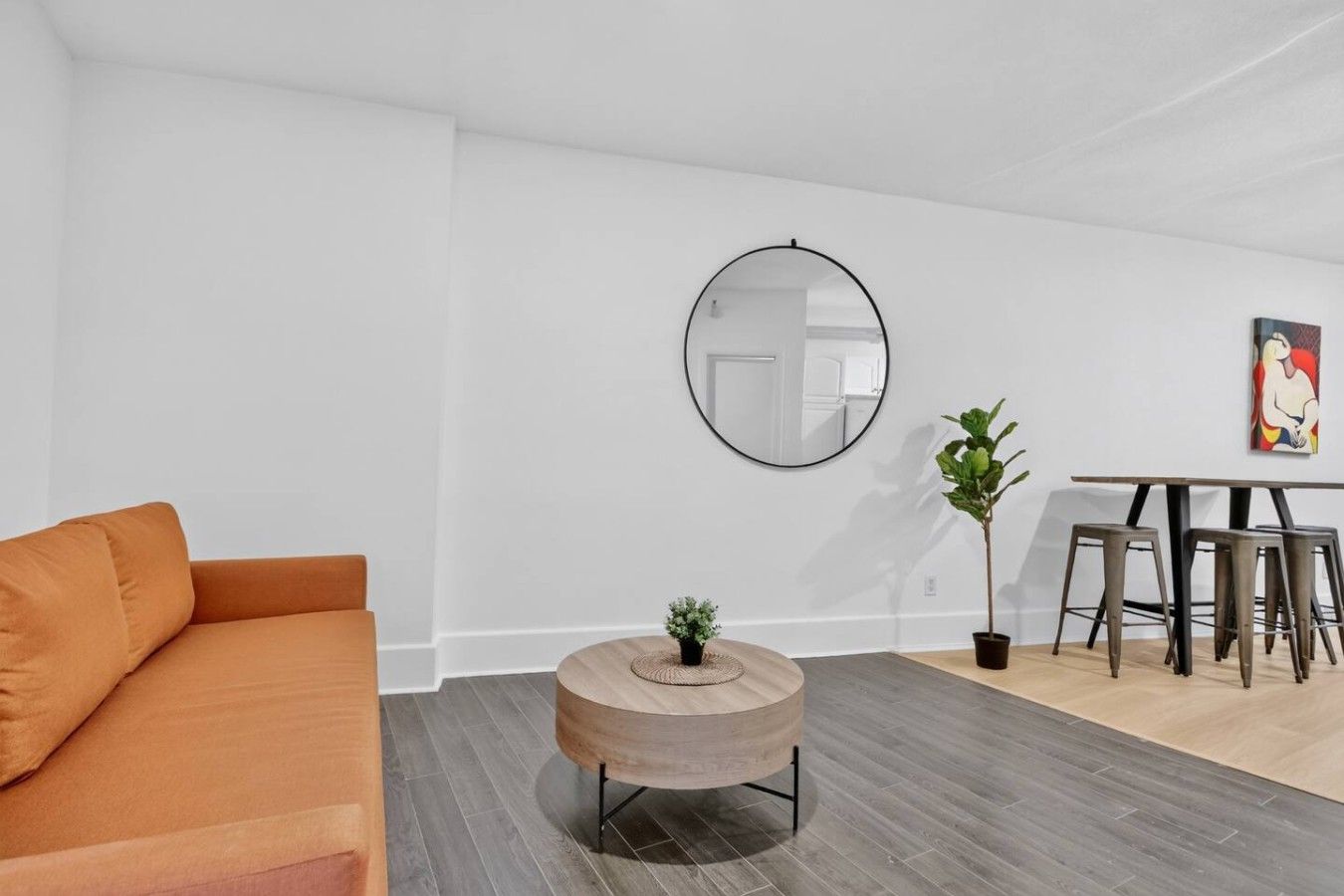 A modern living room with a sofa, a round wooden coffee table with a plant, a dining area with a table and chairs, and a large mirror on the wall.