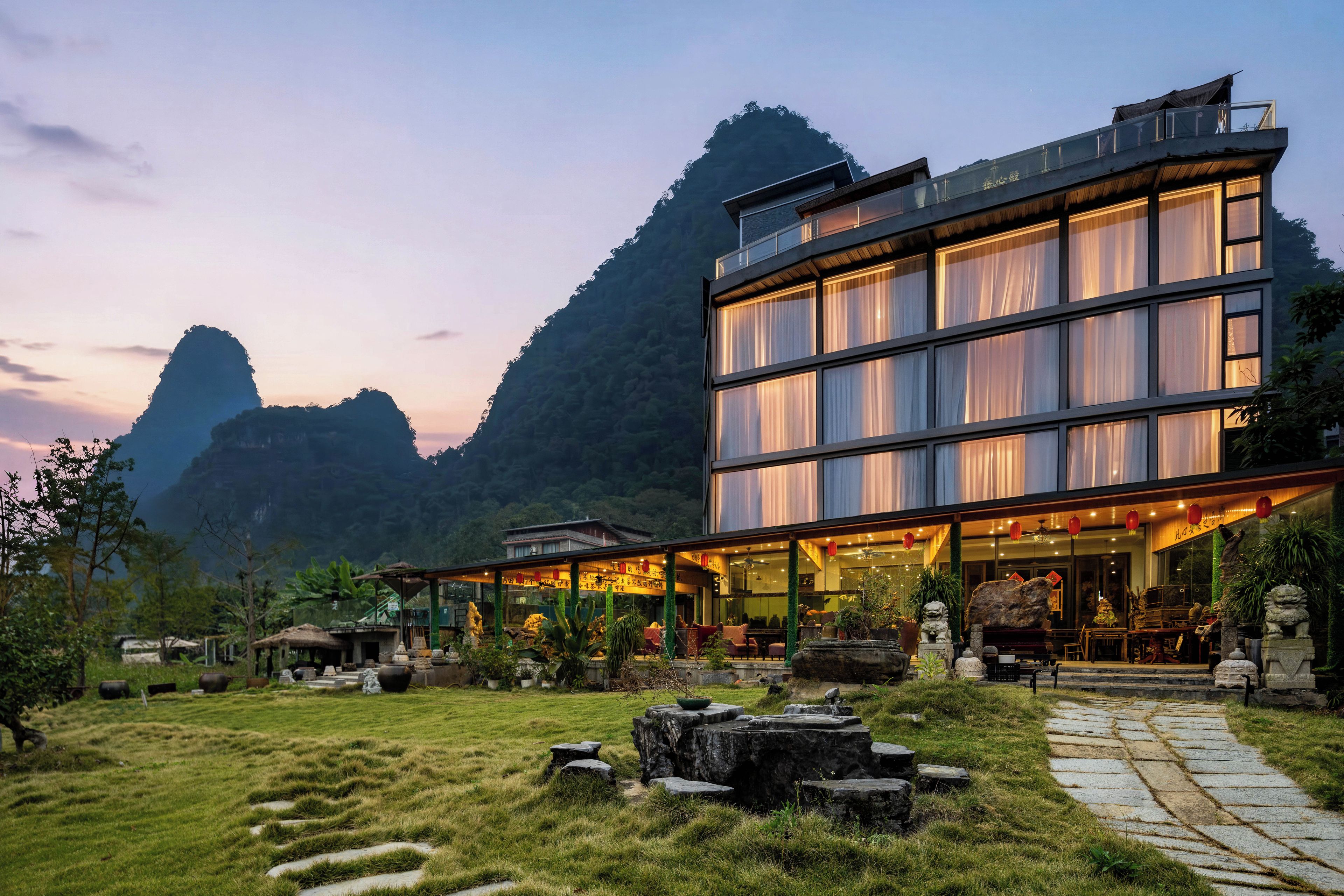 A modern building with a glass facade, surrounded by a landscaped garden with rocks and plants, set against a mountainous backdrop.