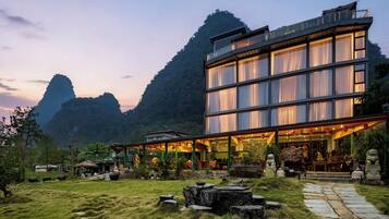 A modern building with a glass facade, surrounded by a landscaped garden with rocks and plants, set against a mountainous backdrop.