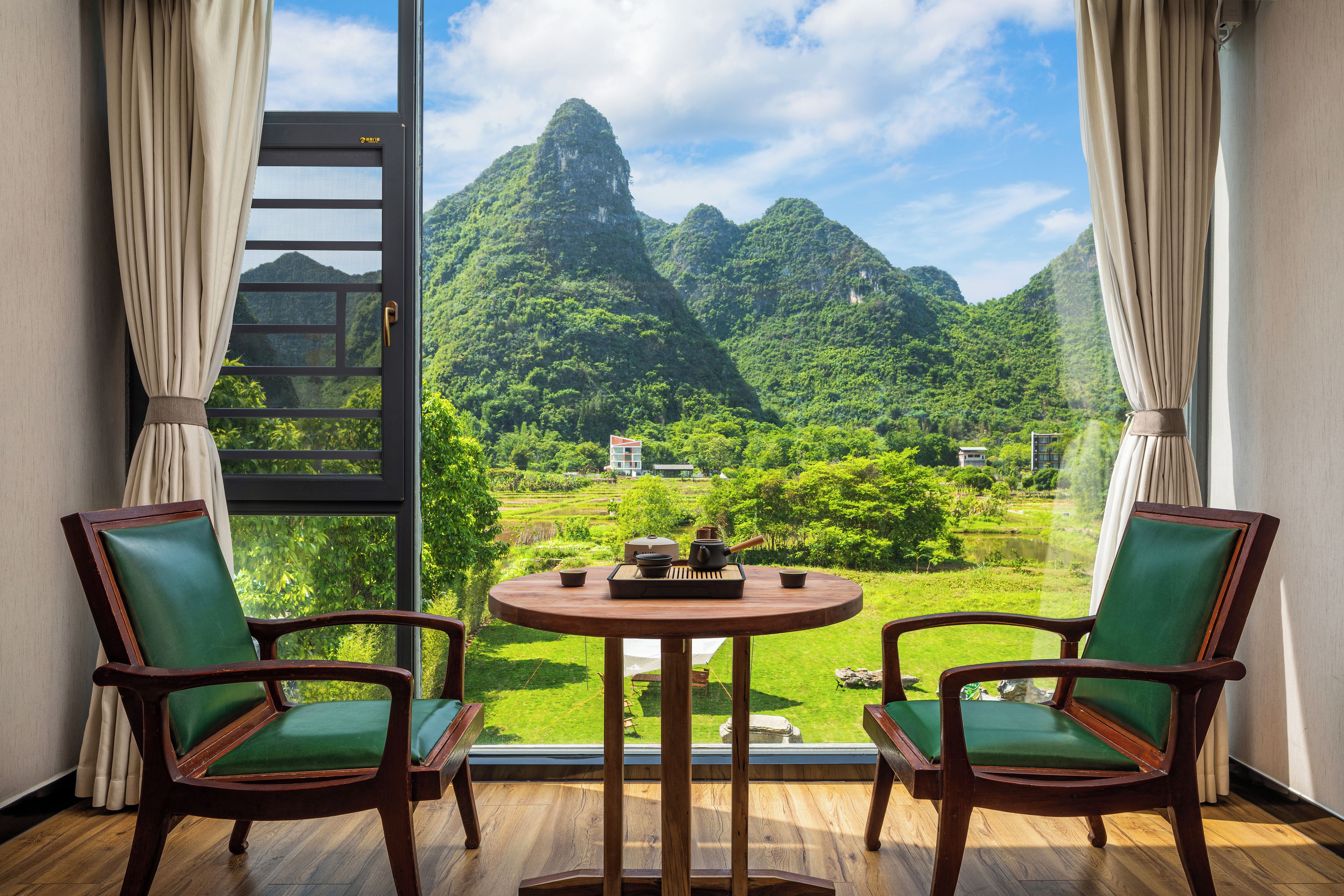A room with a view of a mountainous landscape, two chairs, a table with a teapot and cups, and a ladder.
