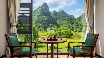A room with a view of a mountainous landscape, two chairs, a table with a teapot and cups, and a ladder.