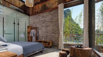 A bedroom with a bed, wooden headboard, a glass wardrobe, a wooden bench, and a view of greenery through a window.