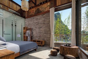 A bedroom with a bed, wooden headboard, a glass wardrobe, a wooden bench, and a view of greenery through a window.