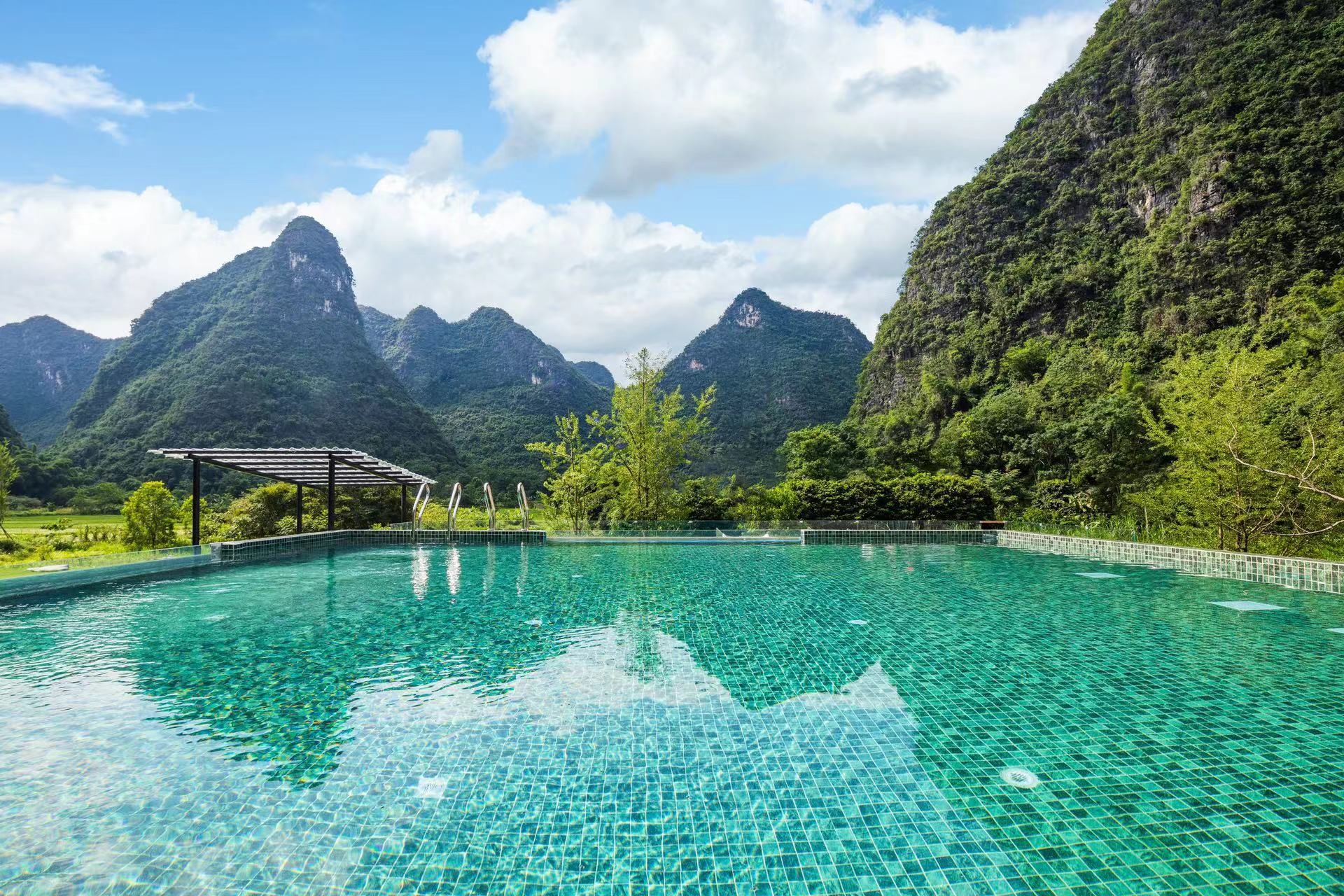 Una piscina cristalina con borde de azulejos, rodeada de exuberante vegetación y montañas al fondo.
