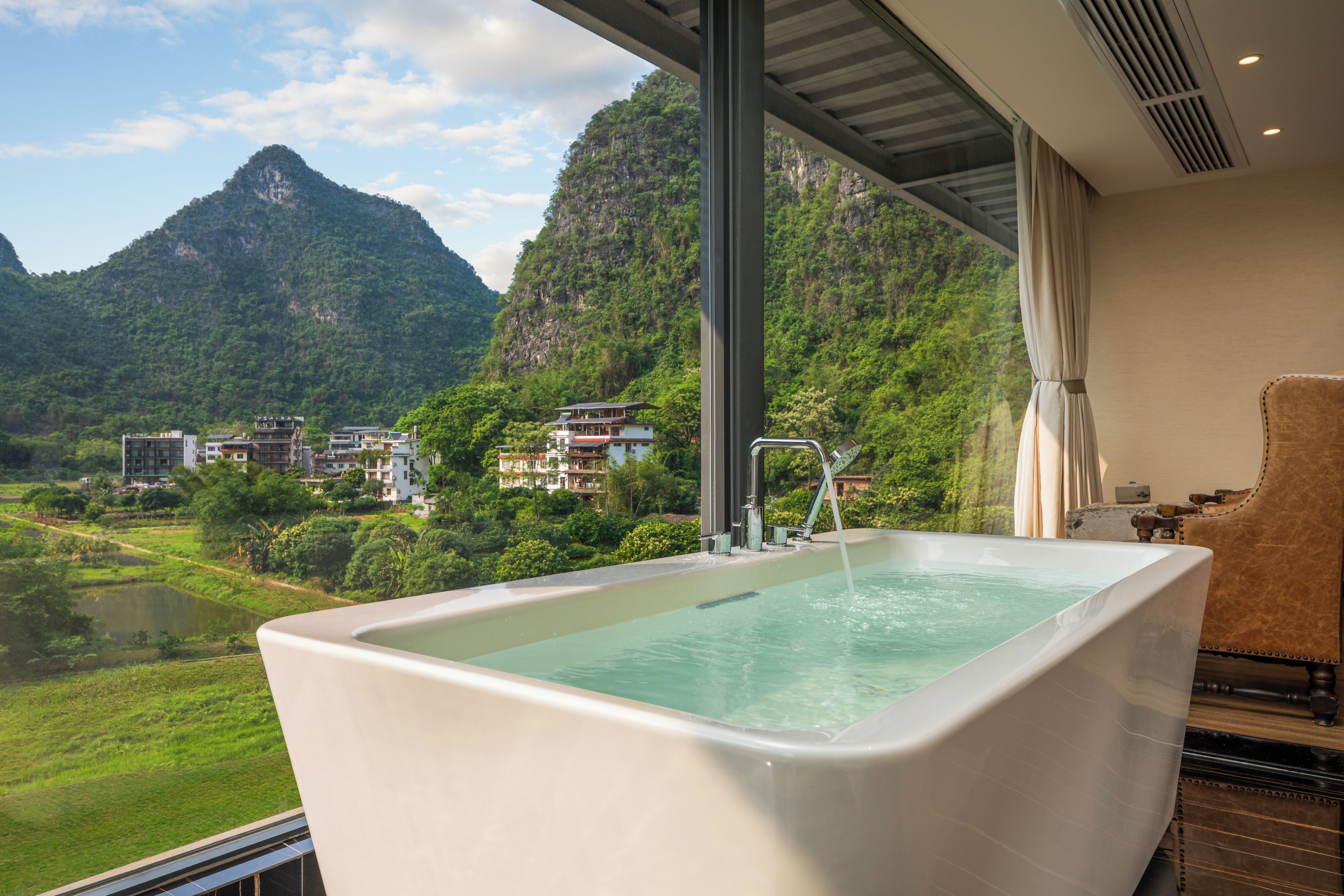 A modern bathroom with a large bathtub, a view of a mountainous landscape, and a glimpse of a hotel building in the background.