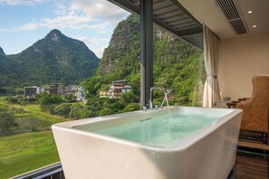 A modern bathroom with a large bathtub, a view of a mountainous landscape, and a glimpse of a hotel building in the background. - Chennianwangshiyisu (Guilin)