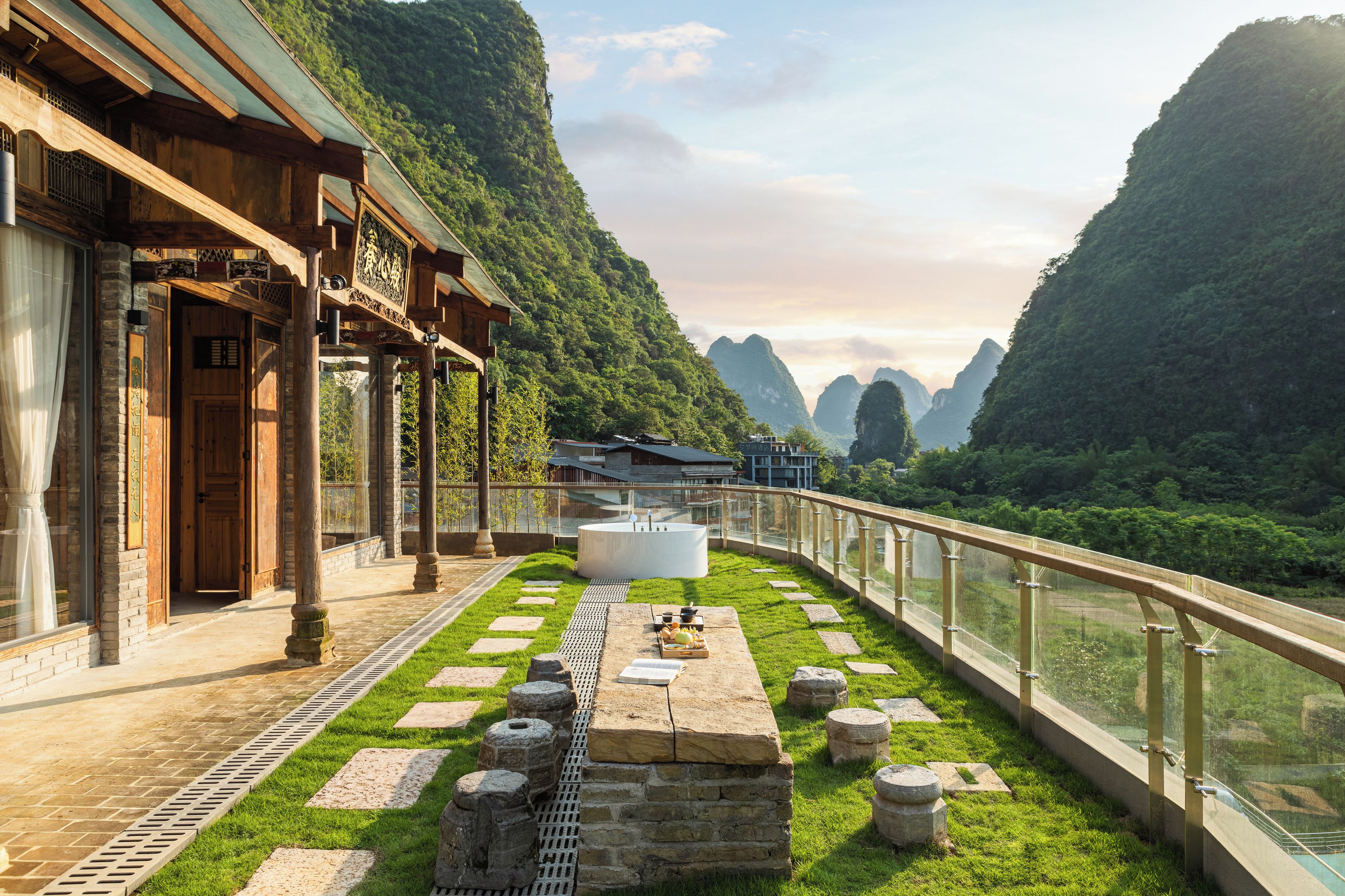 A traditional wooden building with a stone pathway leading to a white circular structure, surrounded by lush greenery and mountains in the background.