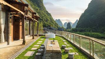 A traditional wooden building with a stone pathway leading to a white circular structure, surrounded by lush greenery and mountains in the background.